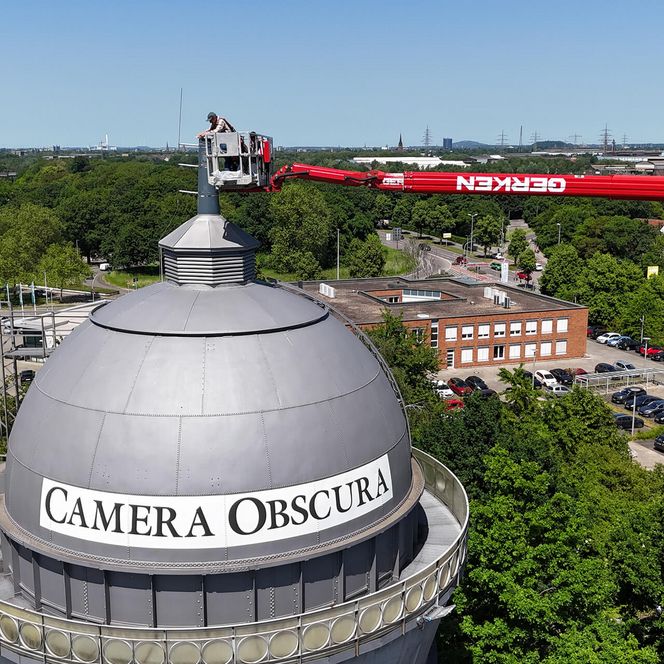 Blick von der Seite auf die Kuppel der Camera Obscura Mülheim: Auf der Plattform eines roten Hubwagens stehen drei Männer und arbeiten an der Spitze des Turms, im Hintergrund erkennt man viele grüne Pflanzen und den Horizont.