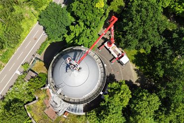 Blick von oben auf die Camera Obscura Mülheim: ein Hubwagen steht neben dem Turm, eine Plattform führt zur Spitze des Turms. Drumherum sind grüne Laubbäume und ein Teil des Radschnellwegs RS1.