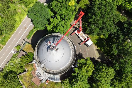 Blick von oben auf die Camera Obscura Mülheim: ein Hubwagen steht neben dem Turm, eine Plattform führt zur Spitze des Turms. Drumherum sind grüne Laubbäume und ein Teil des Radschnellwegs RS1.