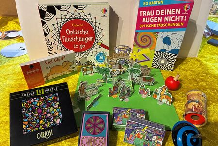 Viele kleine optische Spiele für Kinder, z. B. Puzzle, optische Täuschungen, liegen verteilt auf einer gelbschimmernden Decke
